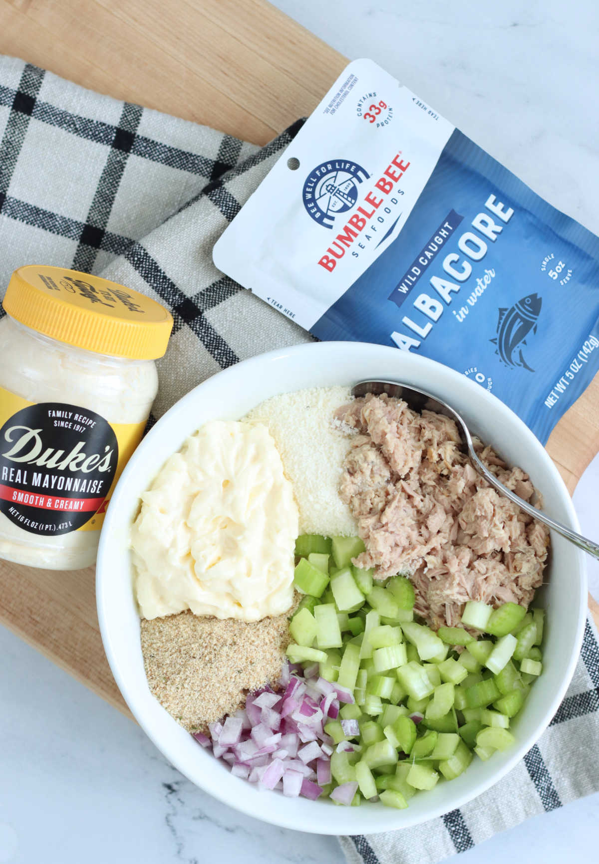 Small white bowl with ingredients for tuna salad, packet of tuna, mayonnaise jar on wooden cutting board.