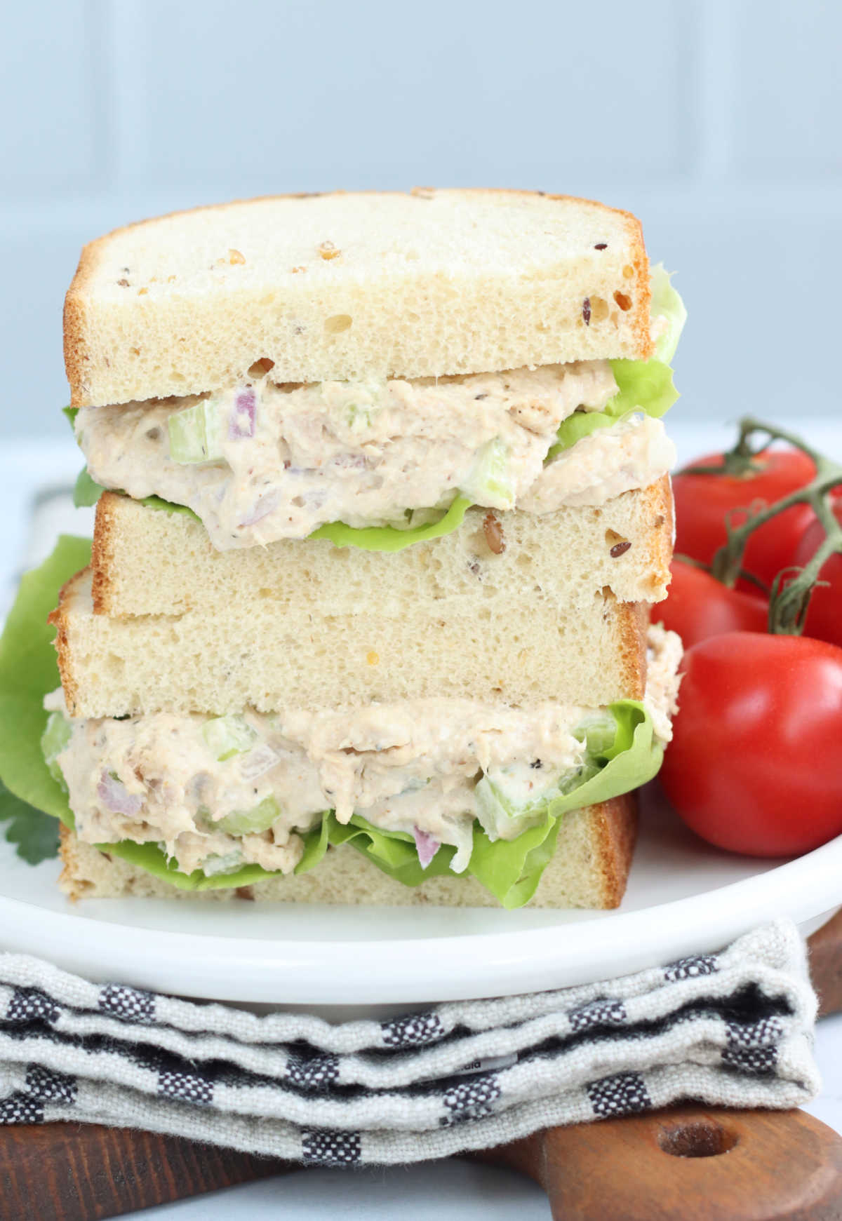 Tuna salad sandwich on whole grain bread with lettuce, stacked on each other on plate, tomatoes to side.