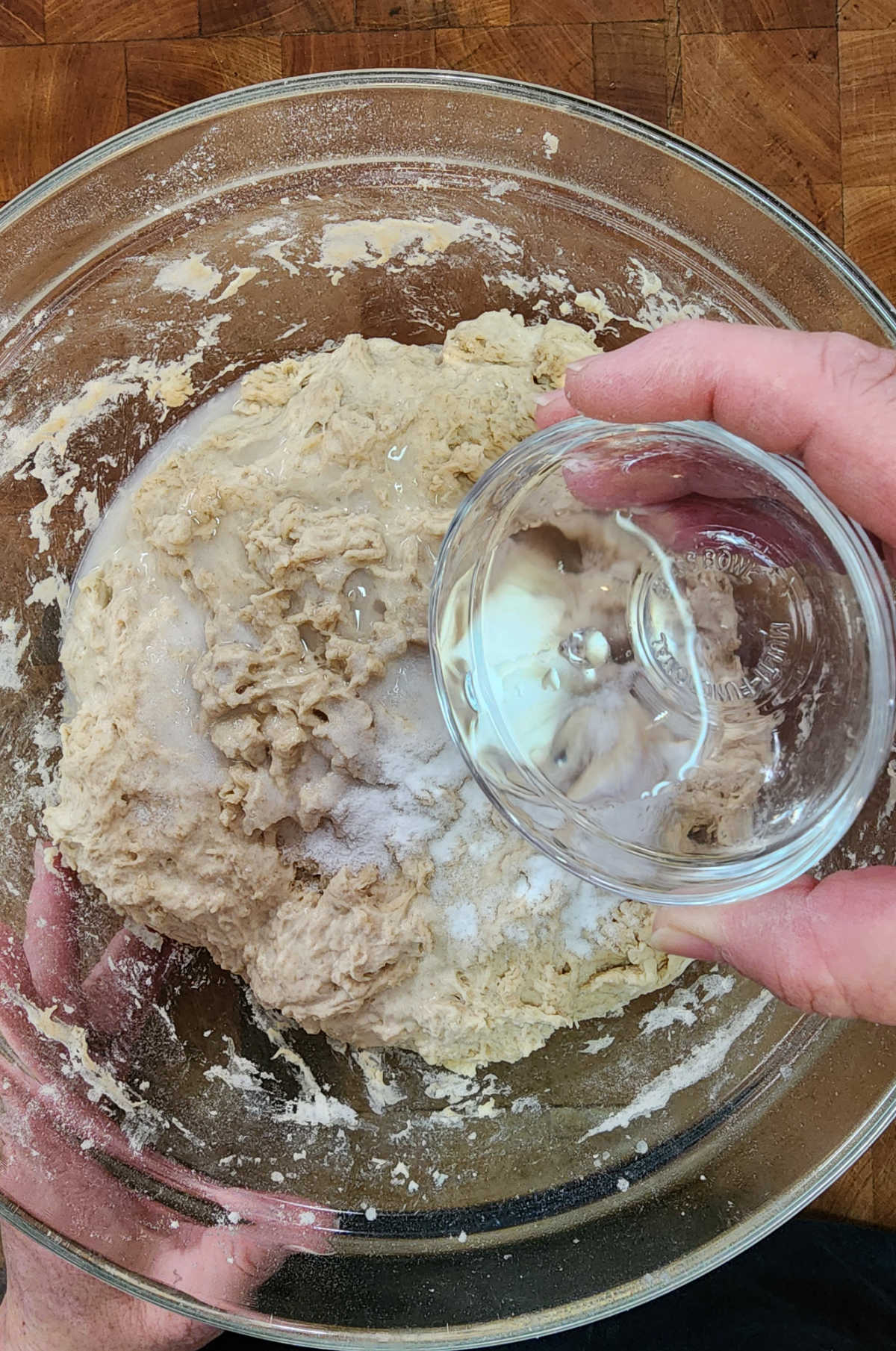 Hand pouring water over sourdough dough in clear glass bowl on butcher block.