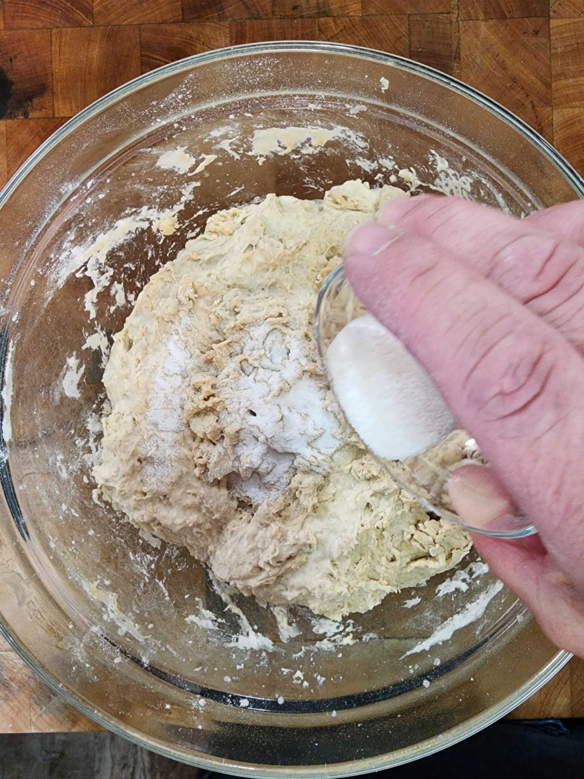 Hand pouring salt over sourdough dough in clear glass bowl on butcher block.