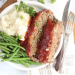 Two slices of meatloaf, mashed potatoes, green beans on small white plate.