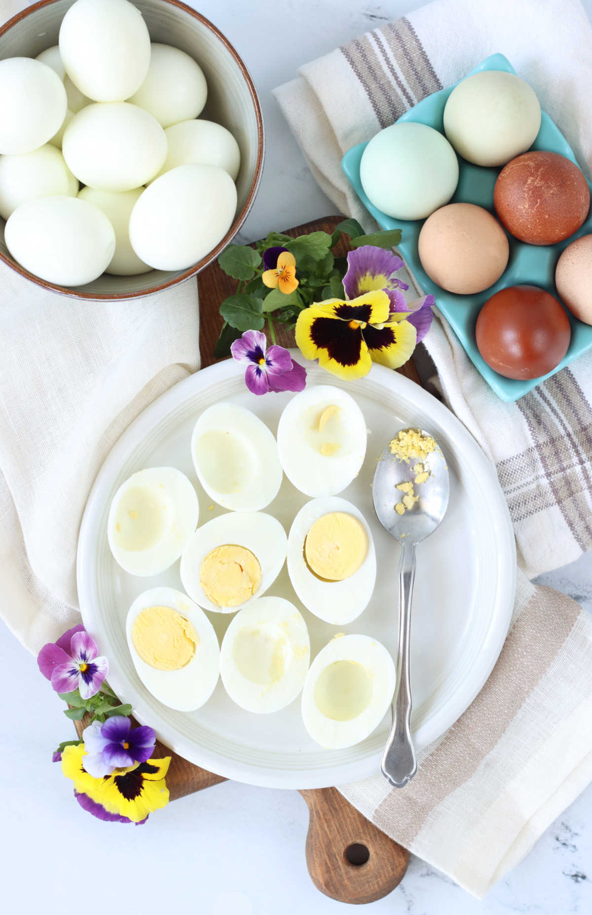 Hard boiled eggs sliced in half on small white plate, eggs in background, pansies.