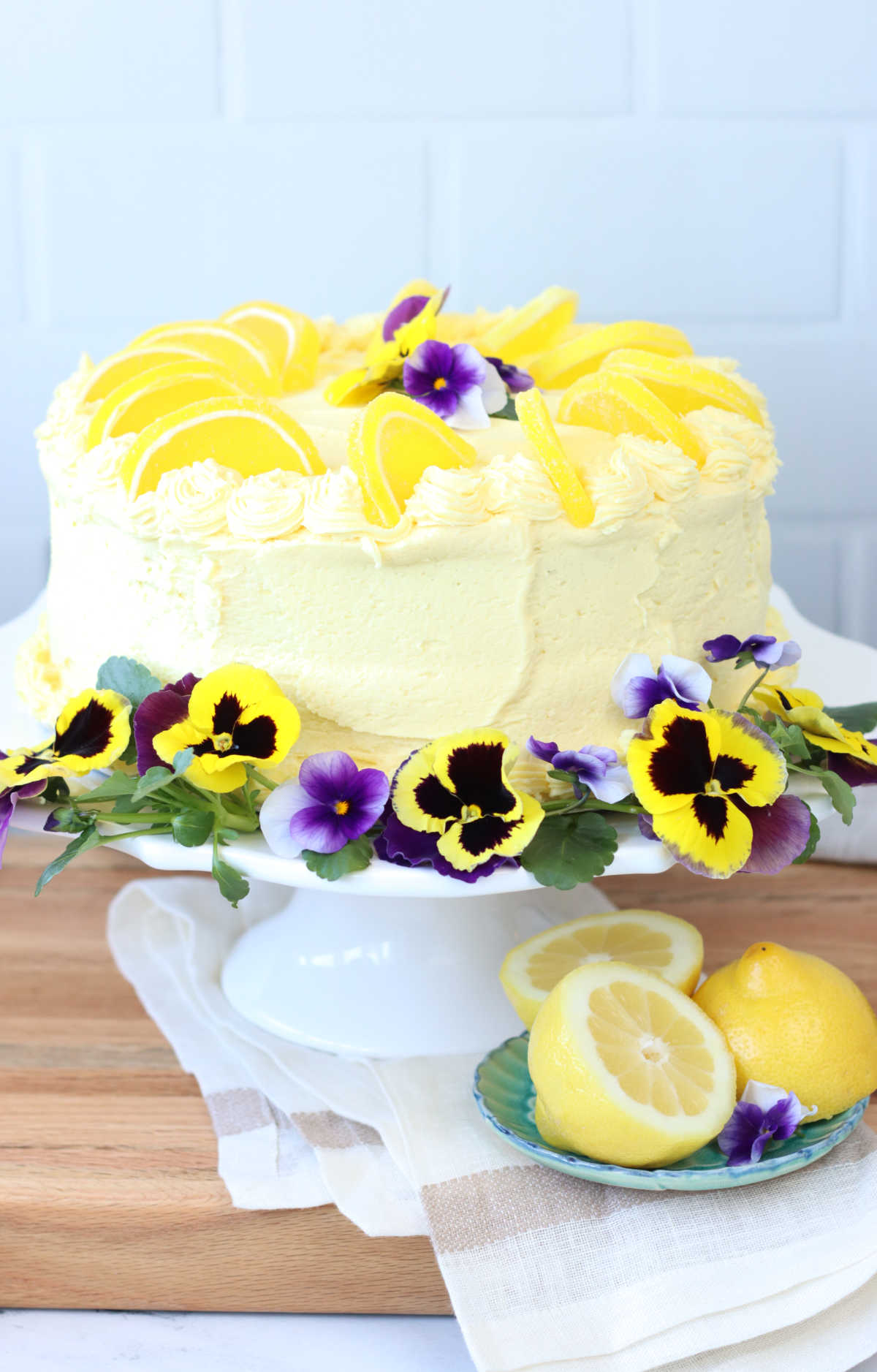 Lemon layer cake on white footed cake dish on wooden cutting board, cake with pansies and violets.
