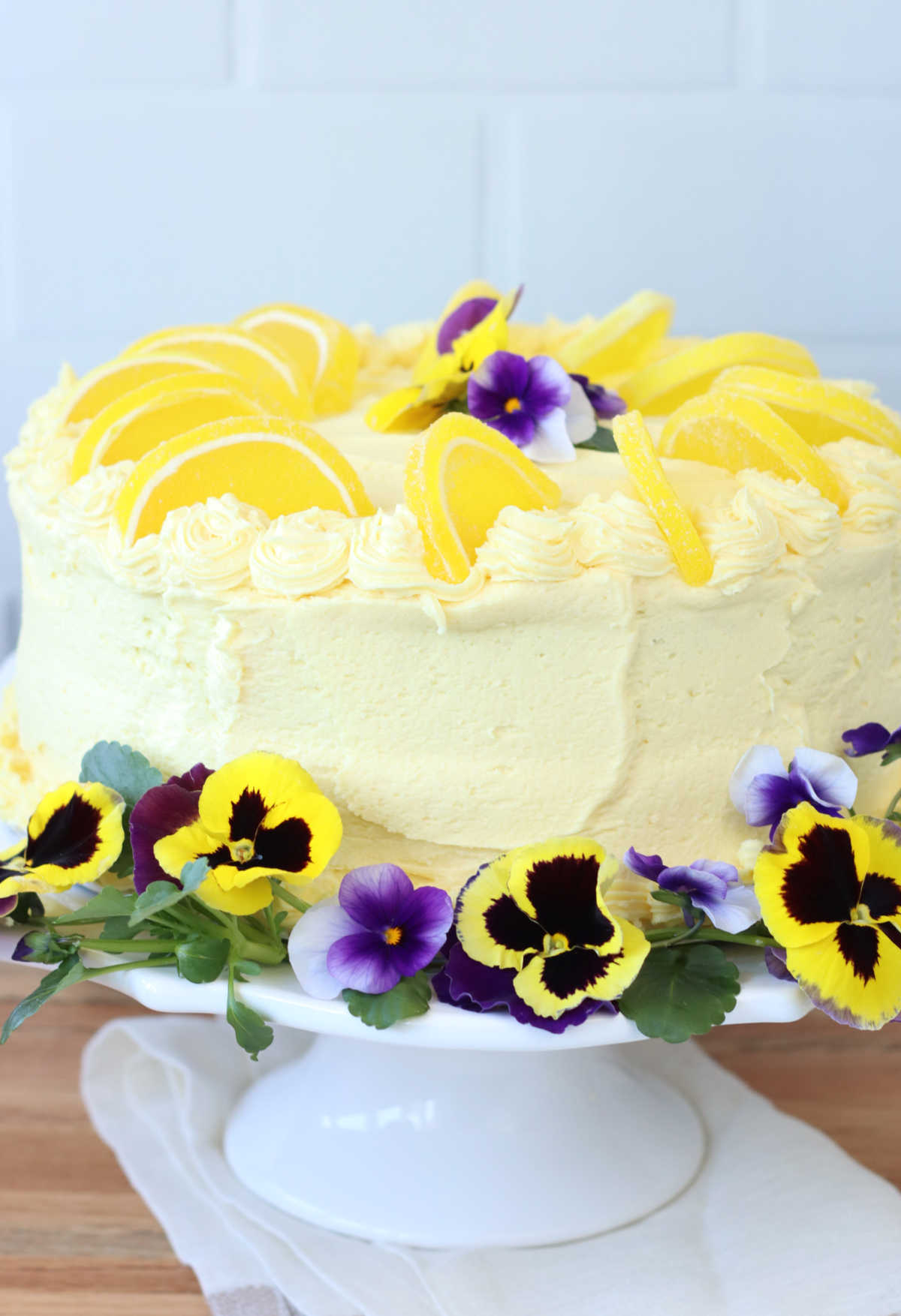 Lemon cake, lemon frosting on white footed cake dish, decorated with lemon candy slices and pansies.