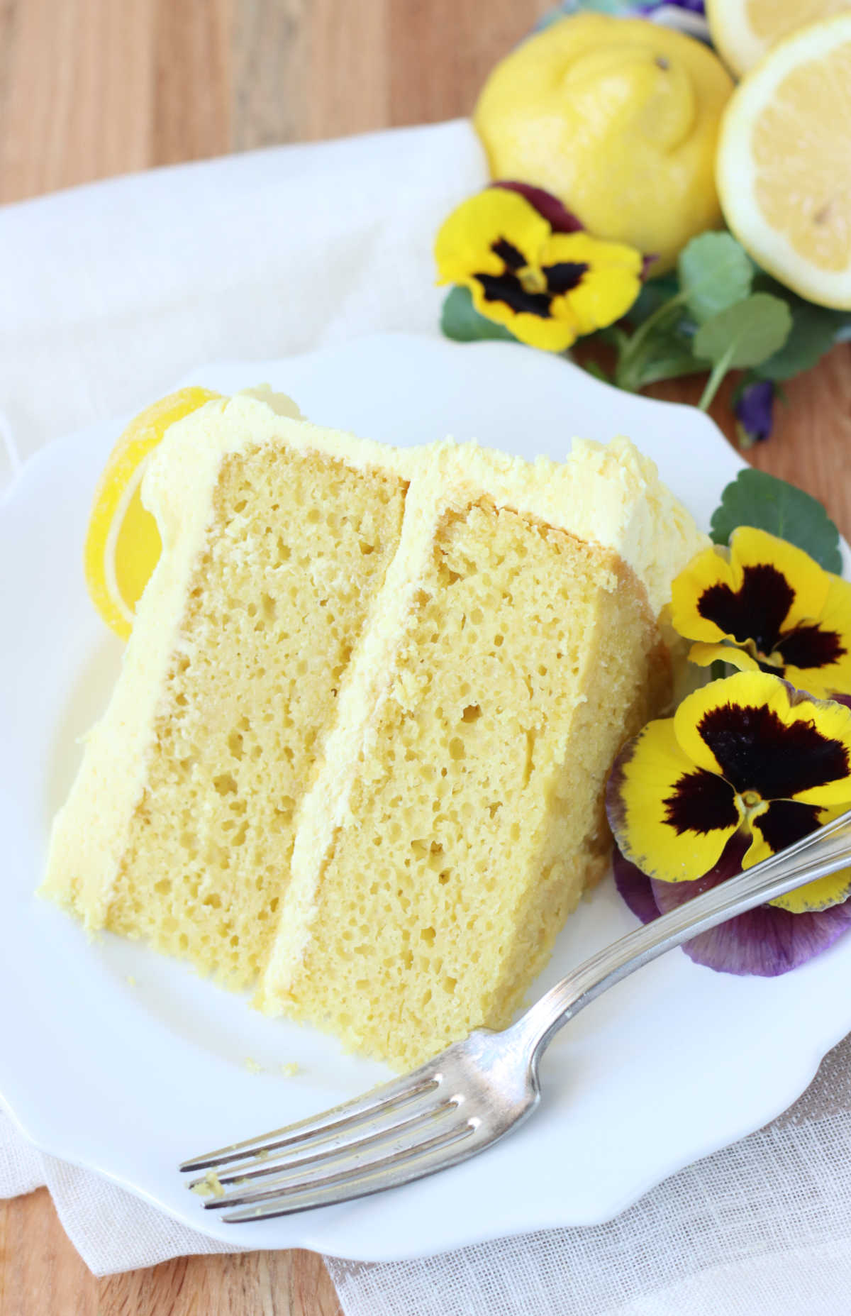 Slice of lemon layer cake on small white plate with fork, fresh lemons in background.