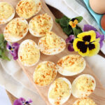 Deviled eggs with paprika on narrow wooden cutting board, pansies around.