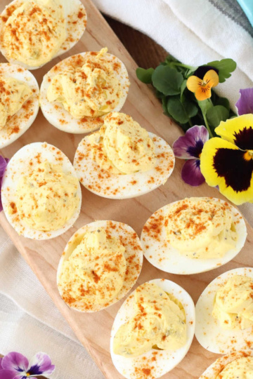 Deviled eggs with paprika on narrow wooden cutting board, pansies around.
