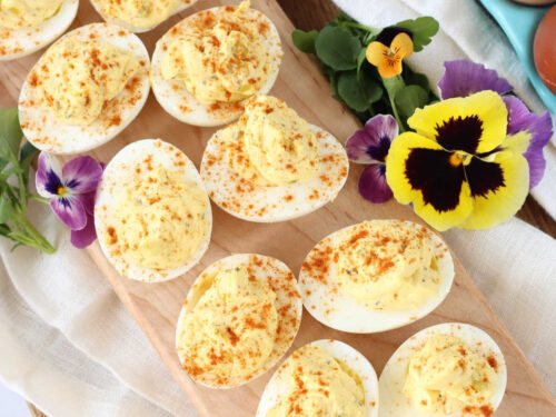 Deviled eggs with paprika on narrow wooden cutting board, pansies around.