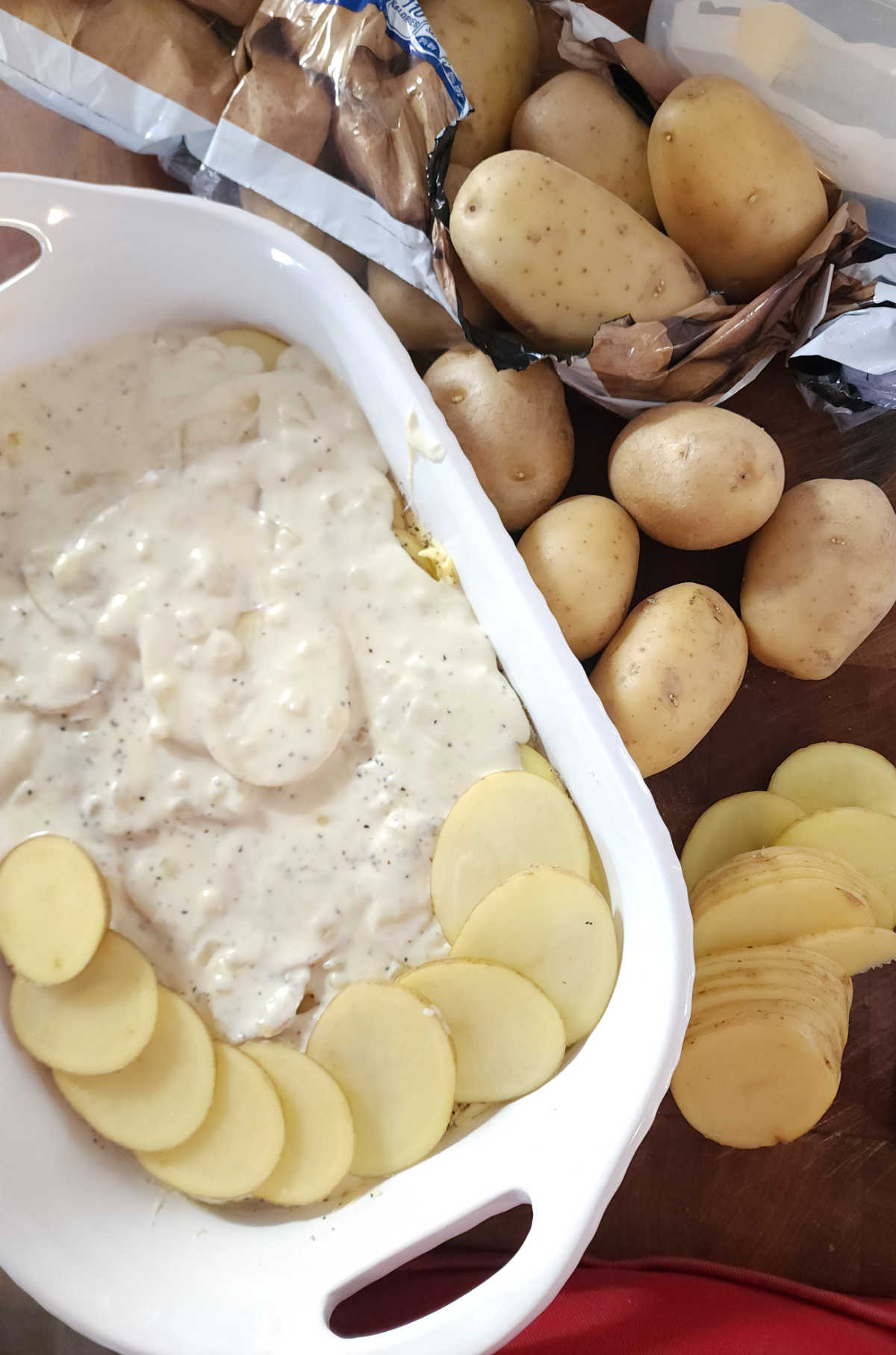 Making au gratin potatoes in white casserole dish on butcher block, slices of potatoes to side.