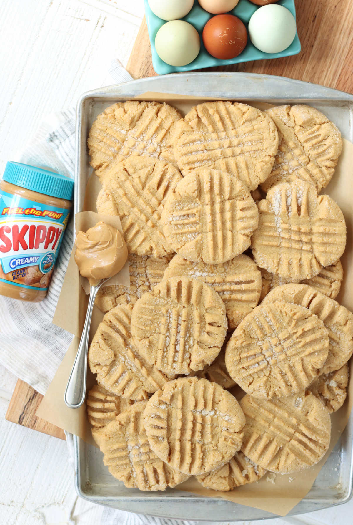 Peanut butter cookies stacked on each other on sheet pan, jar of peanut butter to left, eggs in holder.