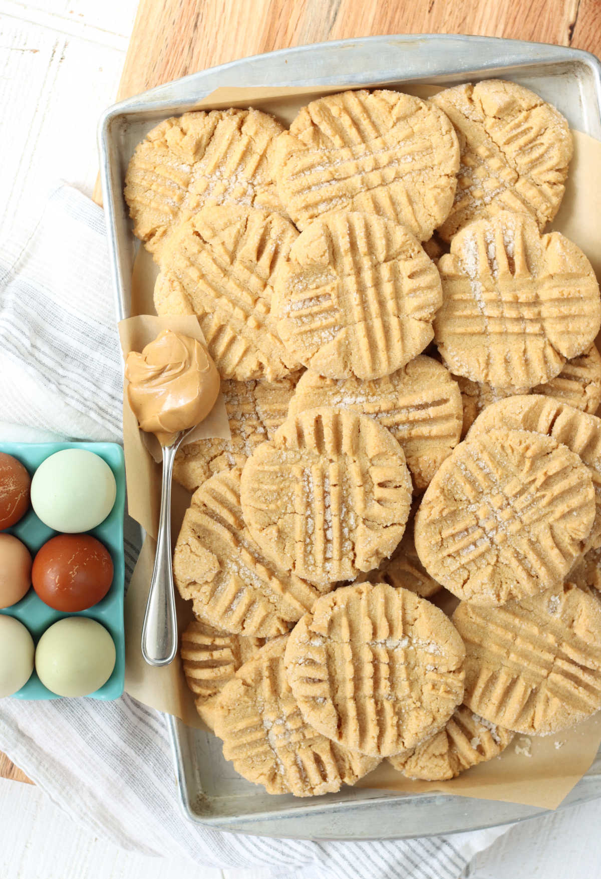 Peanut butter cookies stacked on sheet pan, tablespoon of peanut butter scooped, teal egg holder with colorful eggs to left.