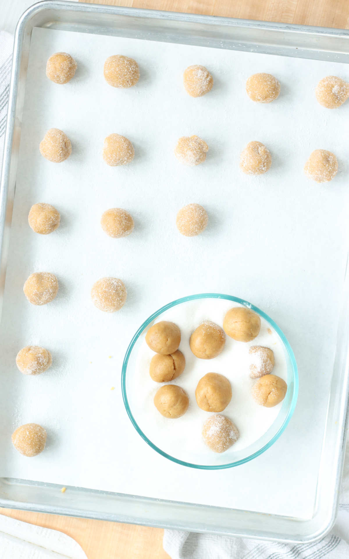 Peanut butter cookie dough rounds on sheet pan, some in clear glass bowl rolled into sugar.