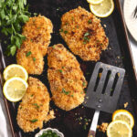 Panko coated chicken breasts on sheet pan, spatula, lemon slices and parsley.