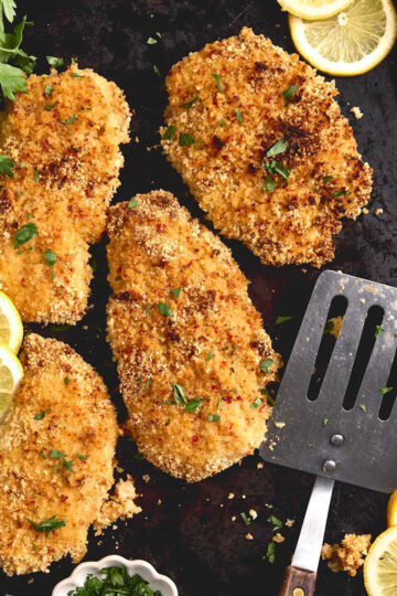 Panko coated chicken breasts on sheet pan, spatula, lemon slices and parsley.