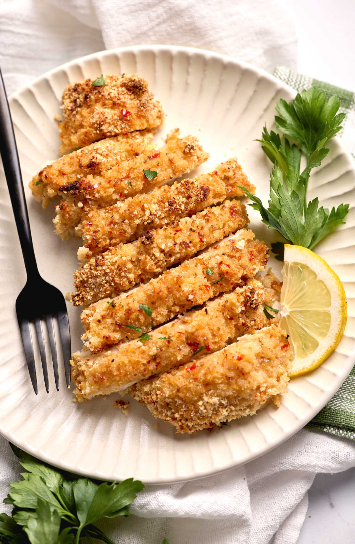 Breaded chicken breast sliced on small white plate with parsley, lemon wedge and fork.