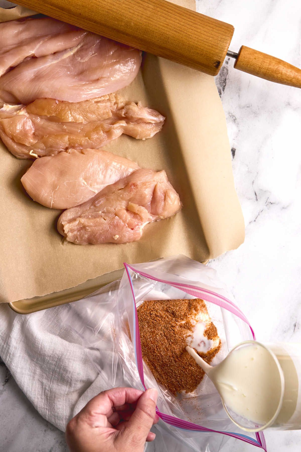 Wooden rolling pin, chicken breasts on sheet pan lined with brown parchment paper, pouring buttermilk into Ziplock bag.