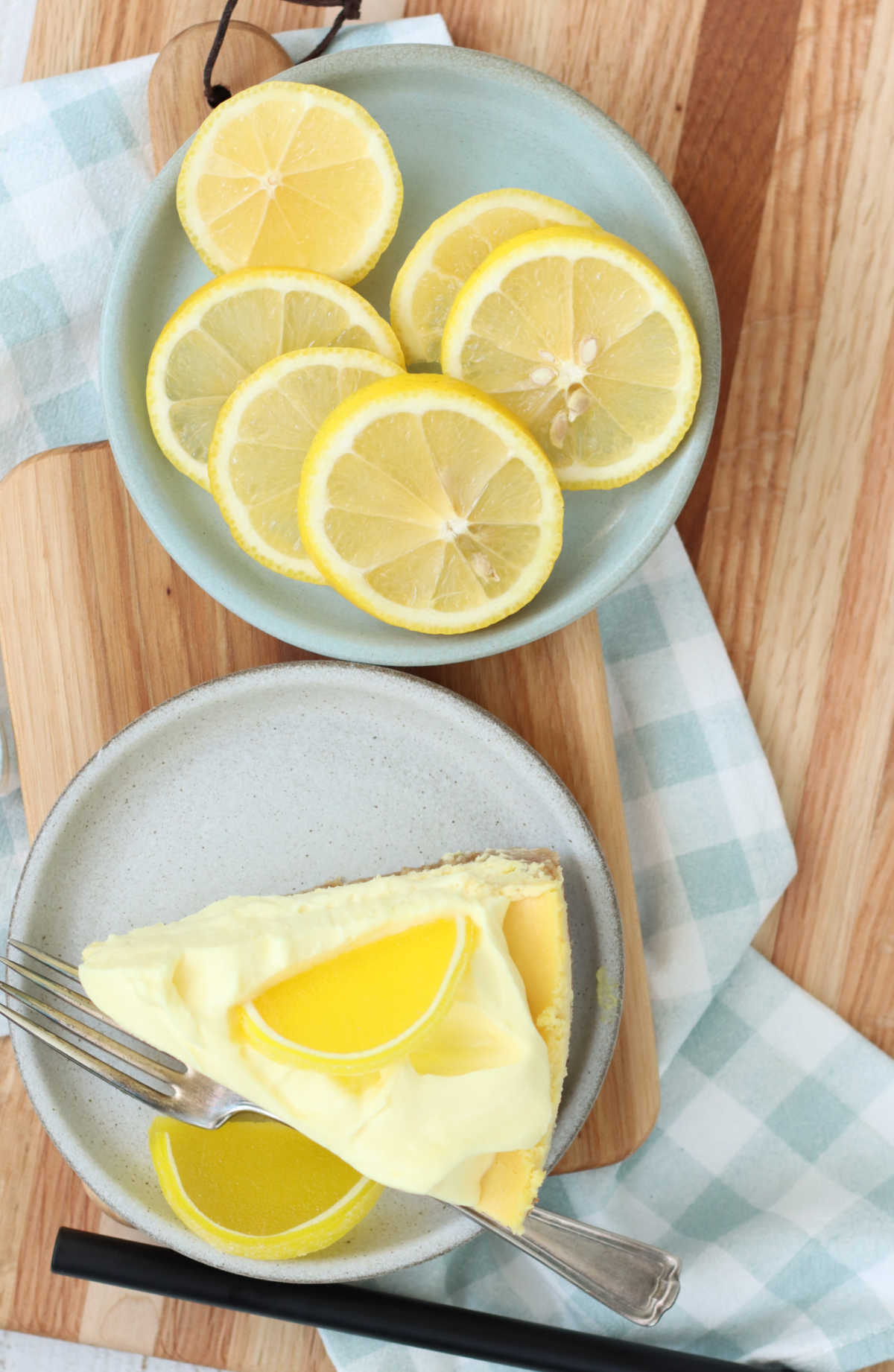 Slice of lemon cheesecake on small plate, plate with slices of lemons on wooden cutting board.