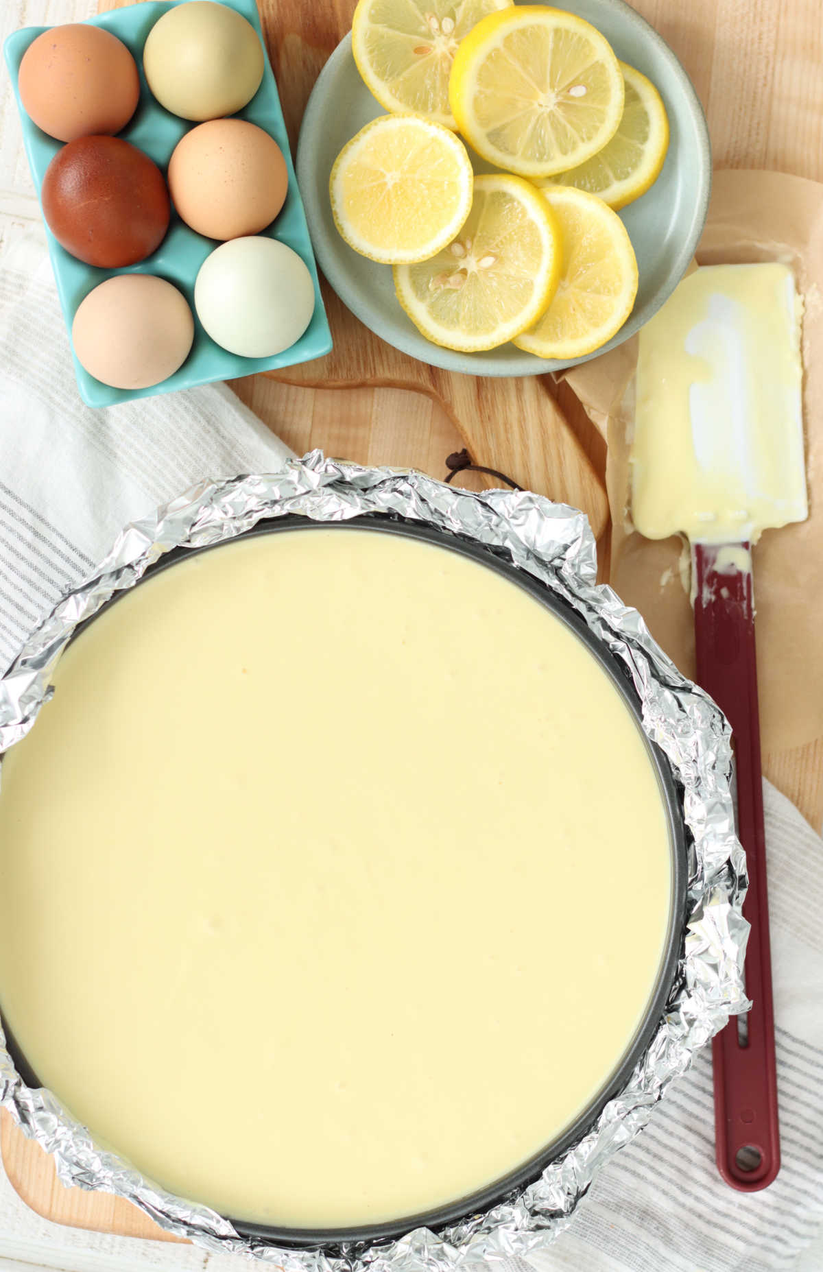 Cheesecake batter in springform pan wrapped with aluminum foil, red handle spatula to right.
