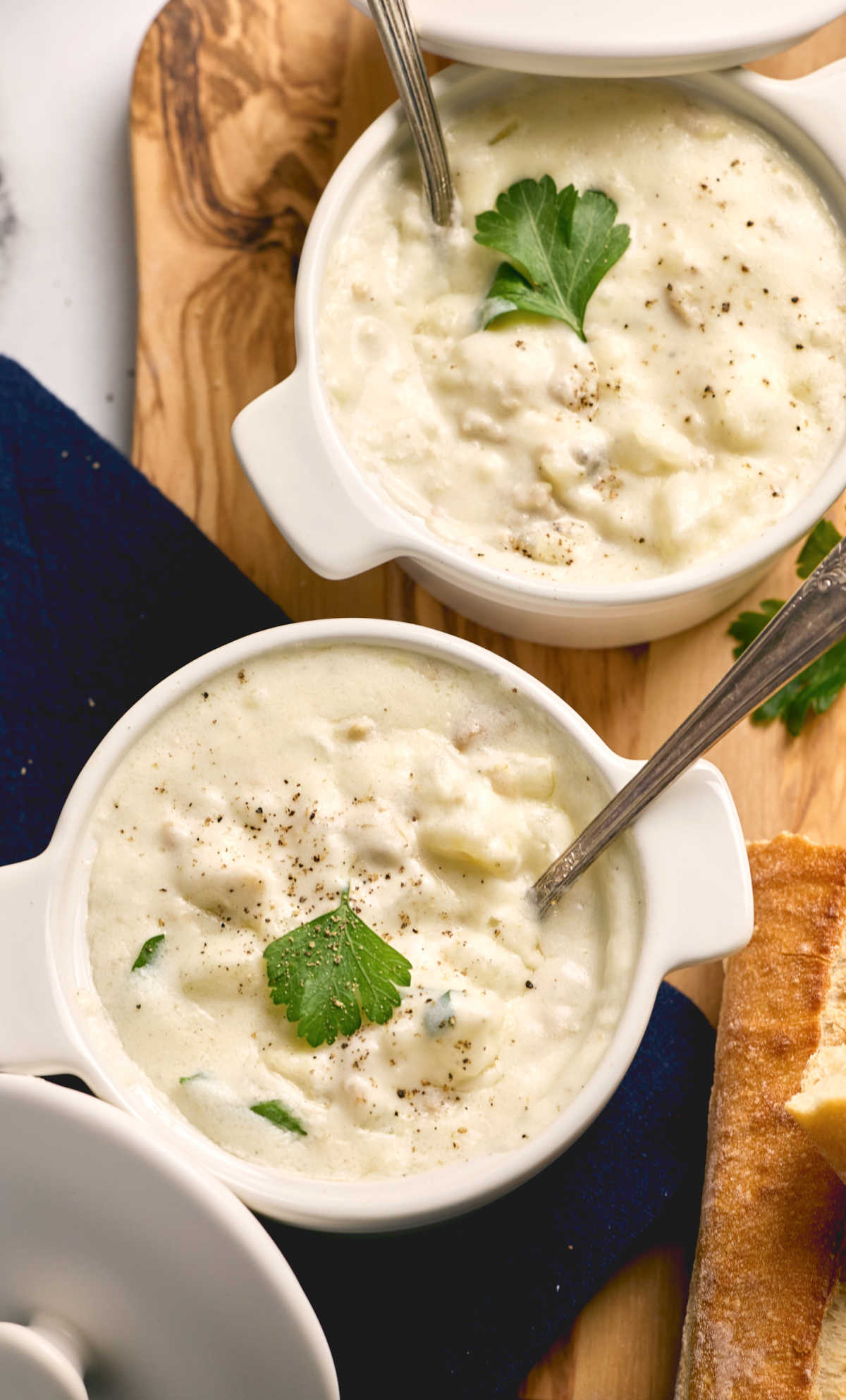 Two white crocks of clam chowder on wooden cutting board.