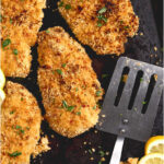 Breaded chicken on sheet pan, fresh parsley, spatula and lemon slices.