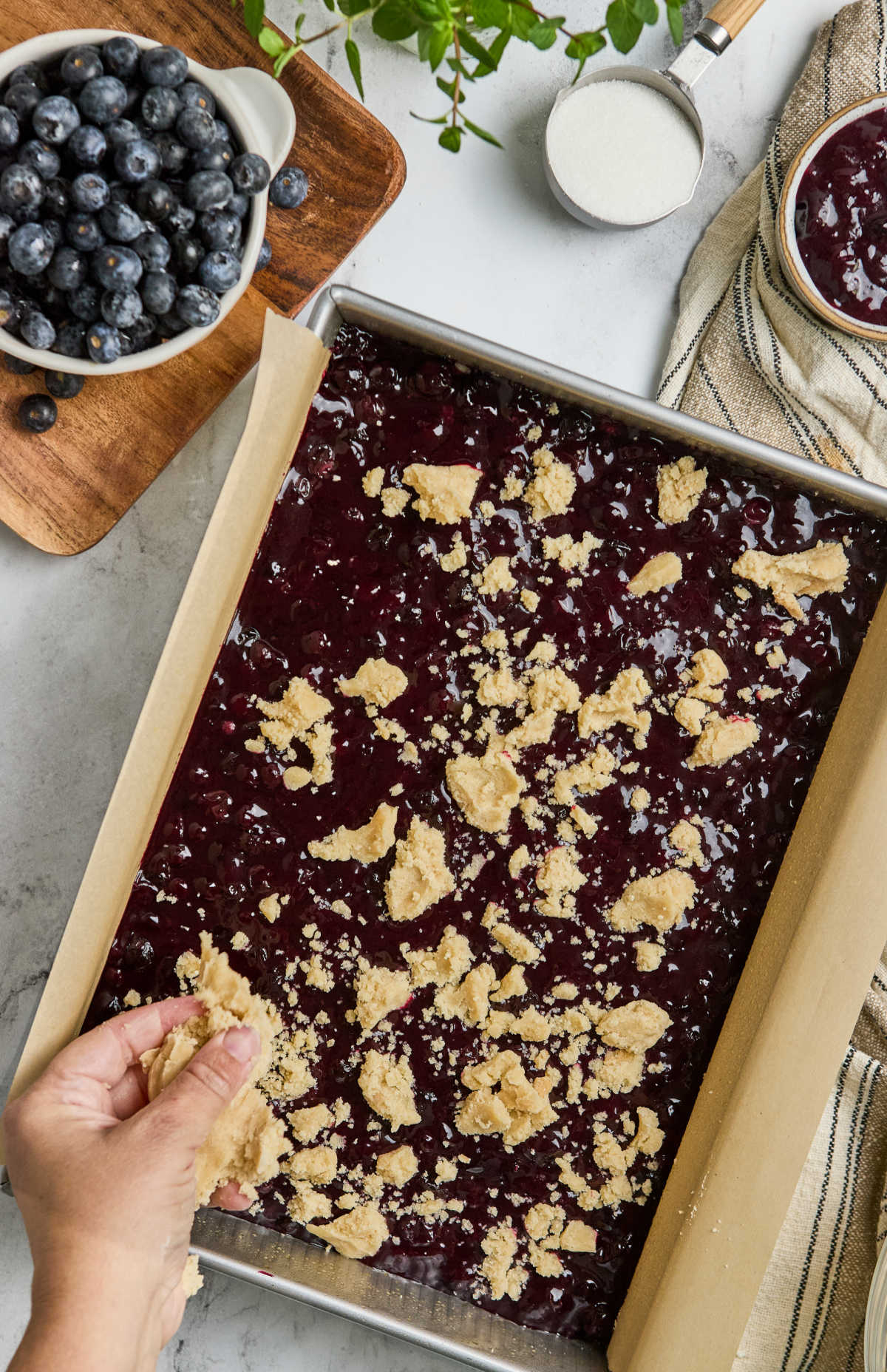 Hand putting crumb on top of blueberry filling in 9x13-inch metal baking pan.