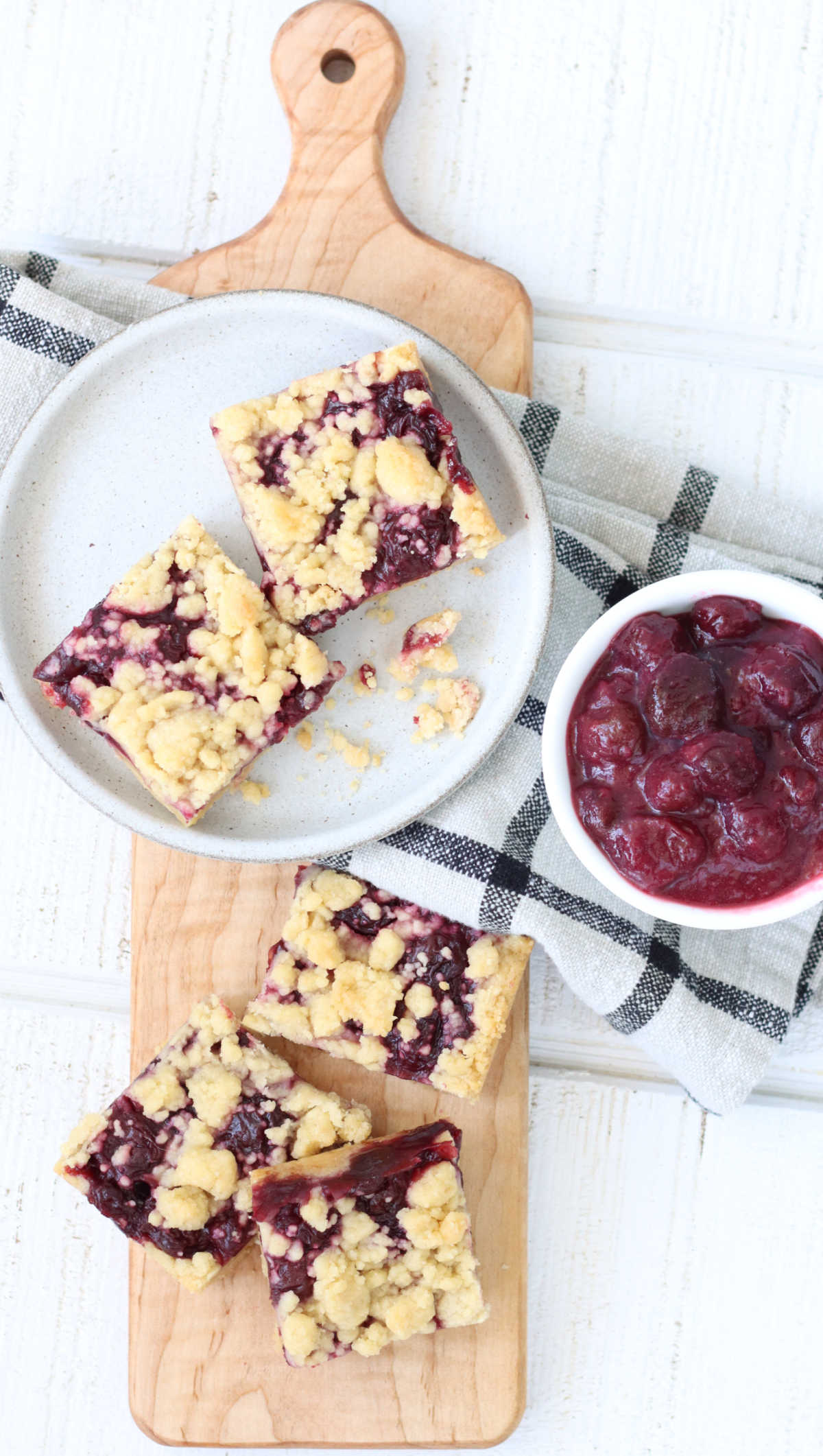Cherry bars cut into squares on small white plate, some on wood cheese board, bowl of cherry pie filling.