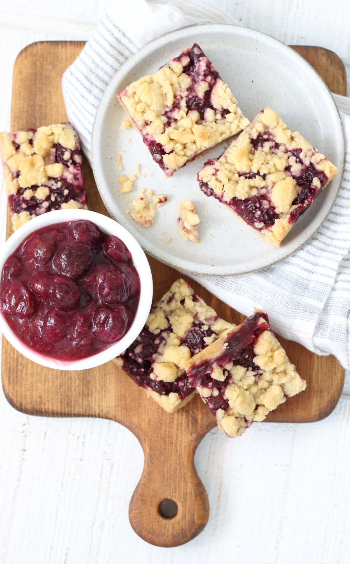 Cherry bars on small white plate, some on wooden cheese board, bowl of cherry filling.
