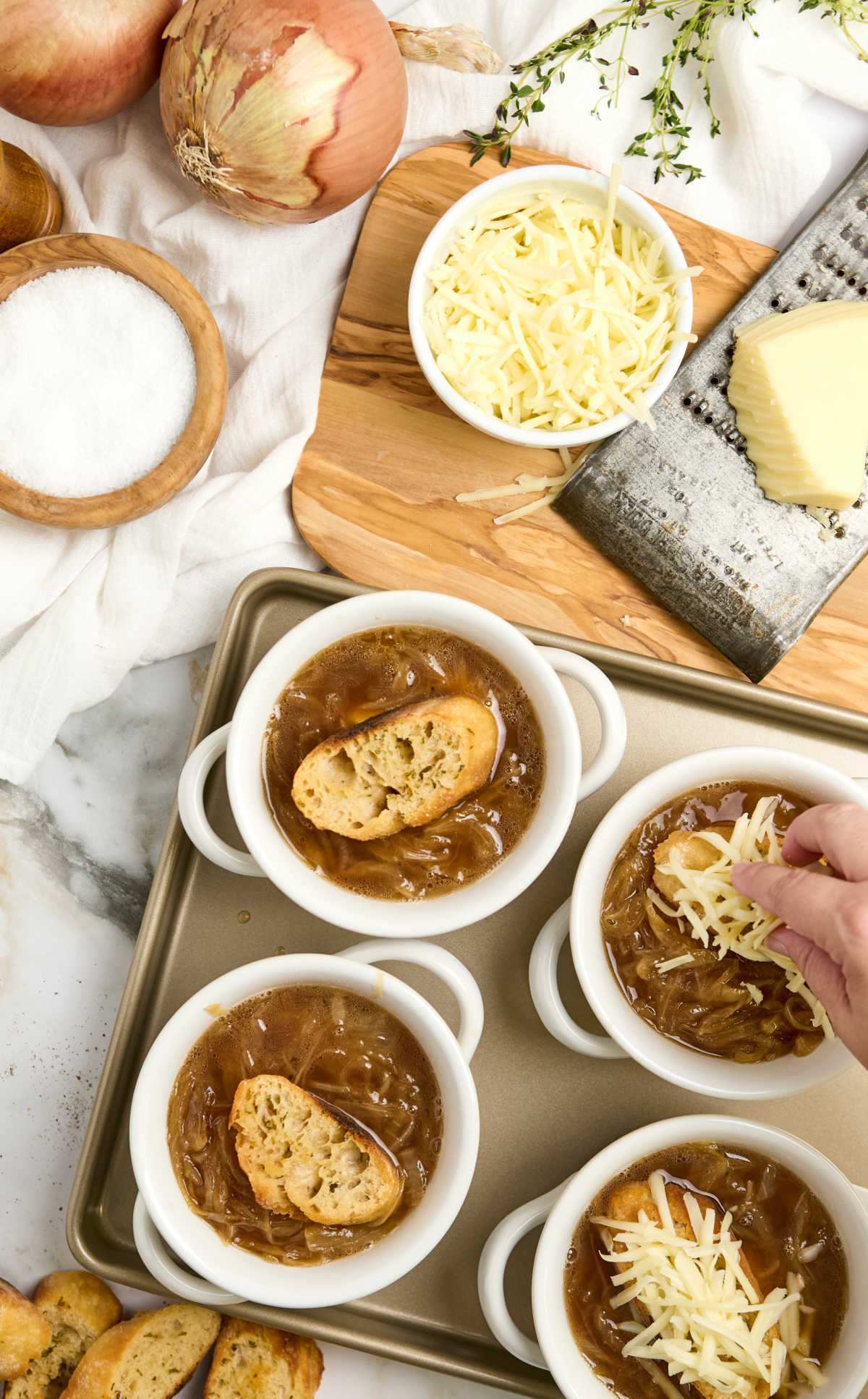 Hand placing grated cheese on top of crocks on French onion soup on sheet pan.