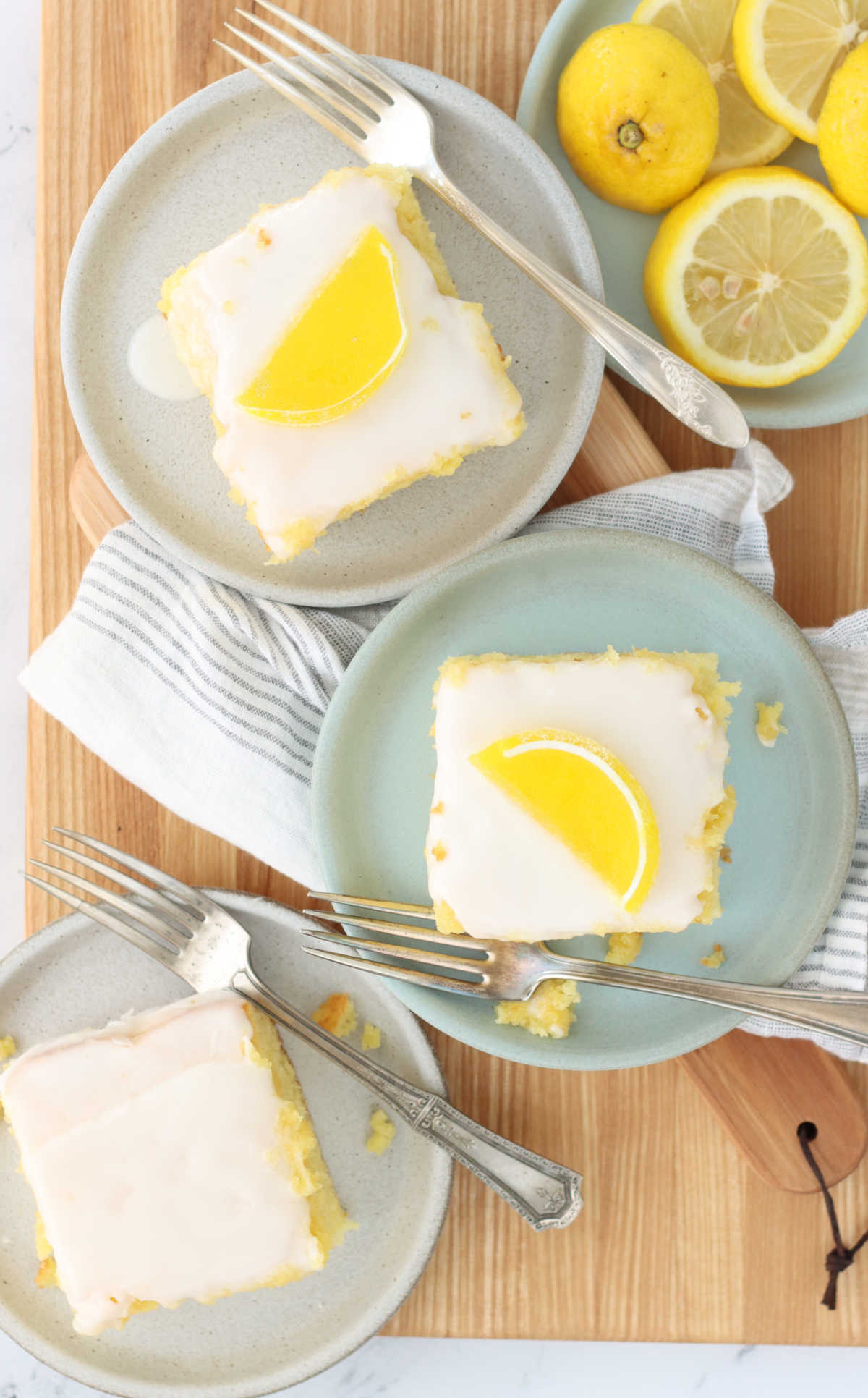 Lemon cake squares on small plates with forks on wooden cutting board, lemon slices on small plate.