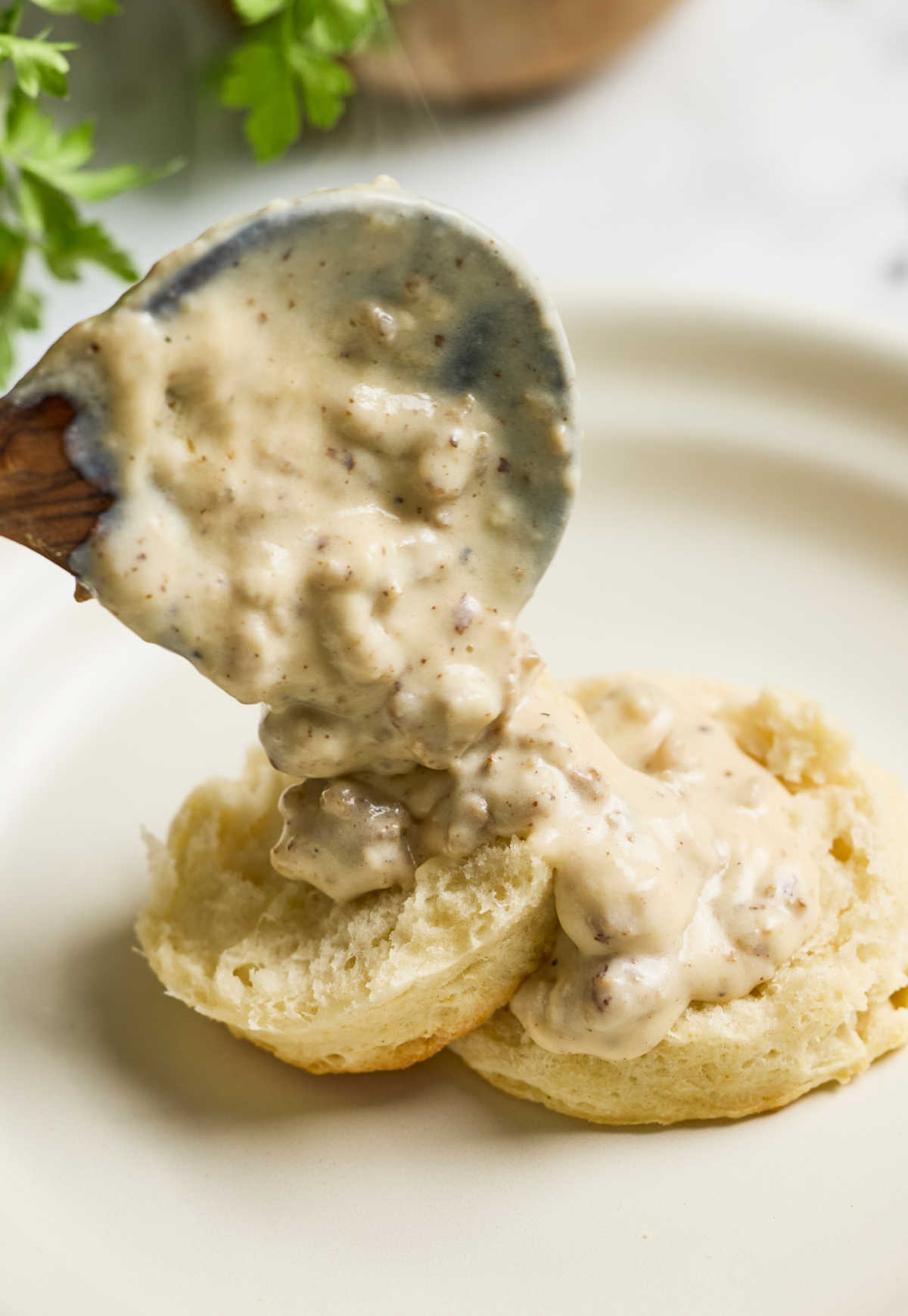 Wooden spoon sausage gravy over biscuits on small white plate.