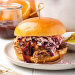 BBQ pork sandwich with coleslaw on small plate, pickles in small bowl.