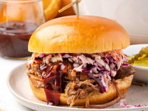 BBQ pork sandwich with coleslaw on small plate, pickles in small bowl.