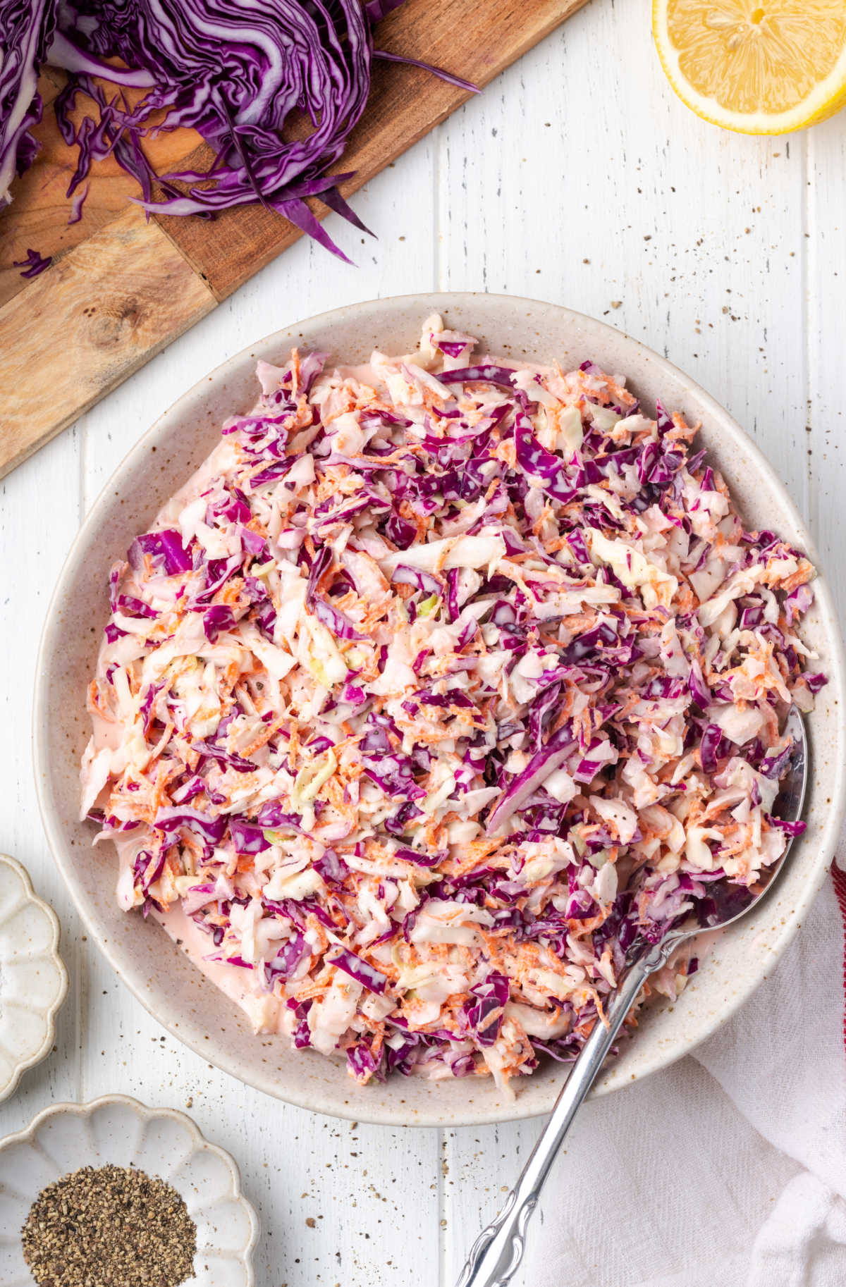 Coleslaw in a white speckled bowl, cabbage in background on wood cutting board.