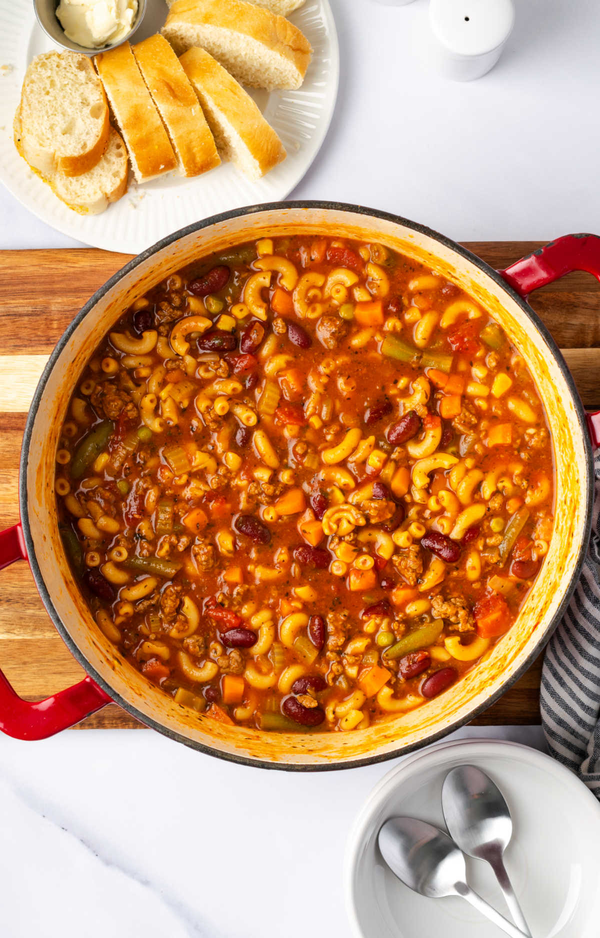 Red Dutch oven with beef vegetable soup on cutting board, slices of bread on white plate.