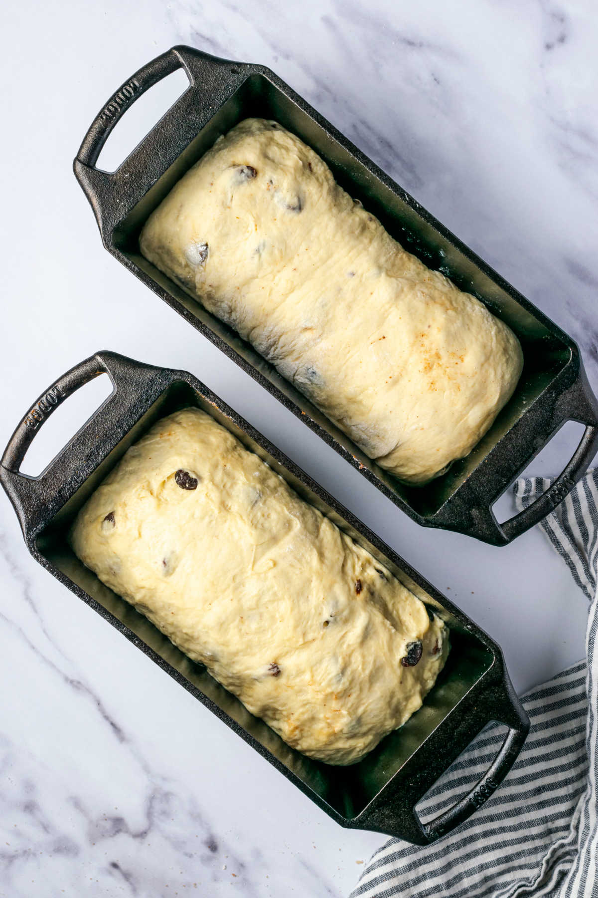 Unbaked cinnamon raisin bread in cast iron loaf pans on white marble.