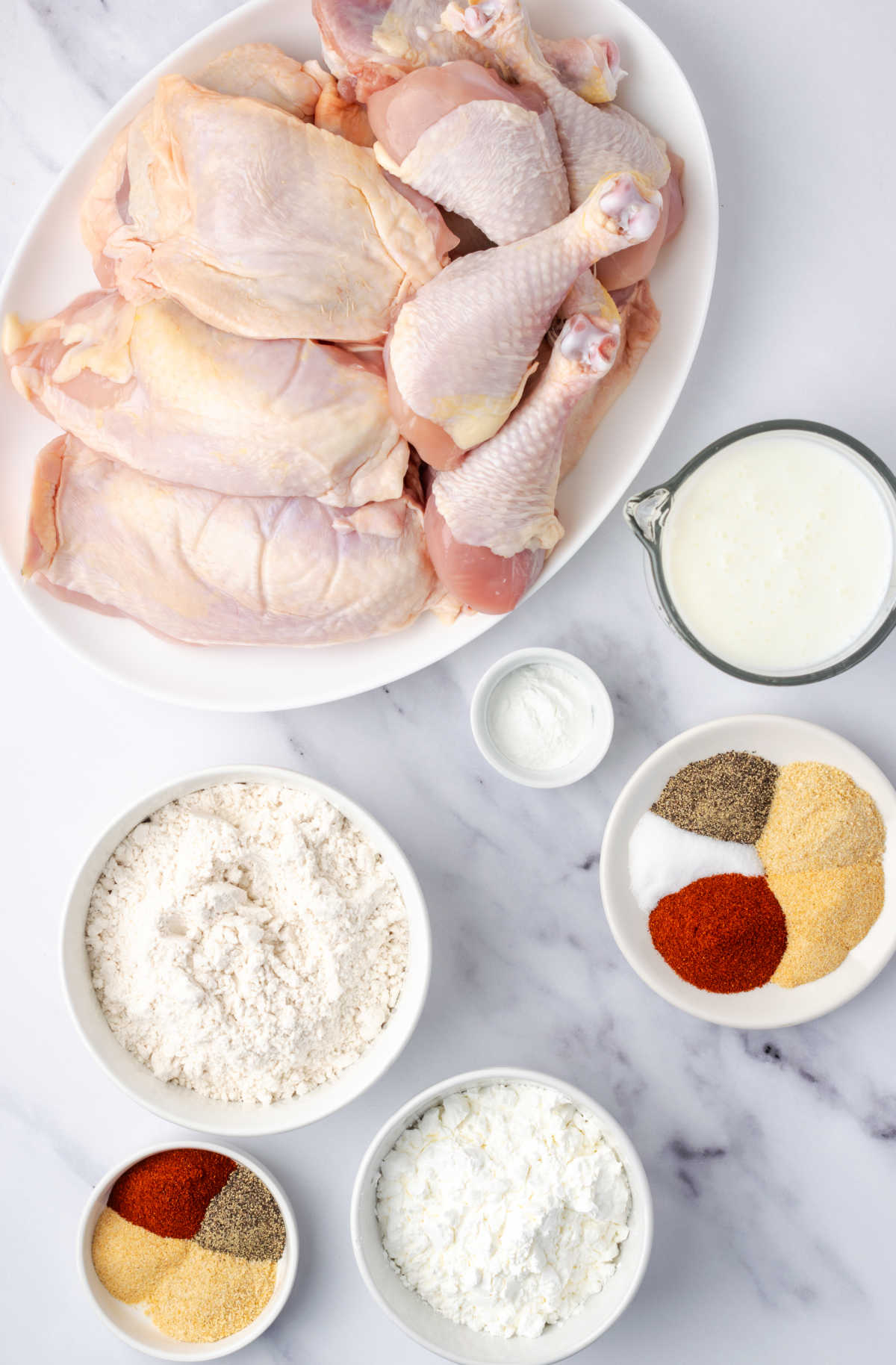 Chicken pieces on oval plate, spices and buttermilk is small bowls on white marble.
