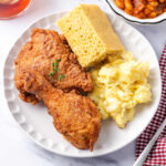 Buttermilk fried chicken on small white plate with cornbread and potato salad.