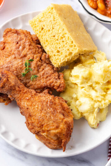 Buttermilk fried chicken on small white plate with cornbread and potato salad.