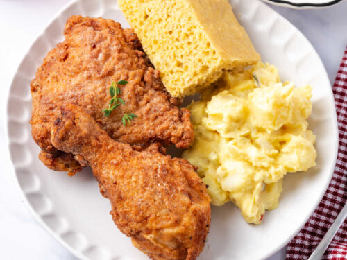 Buttermilk fried chicken on small white plate with cornbread and potato salad.