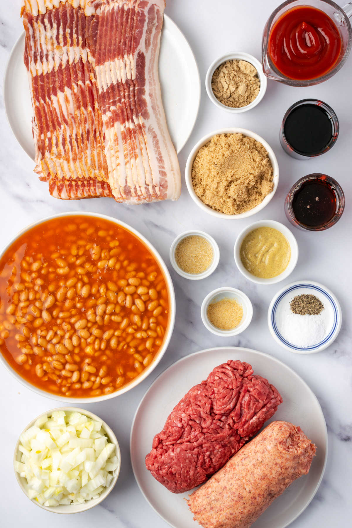 Ingredients for cowboy baked beans in small white bowls on white marble.