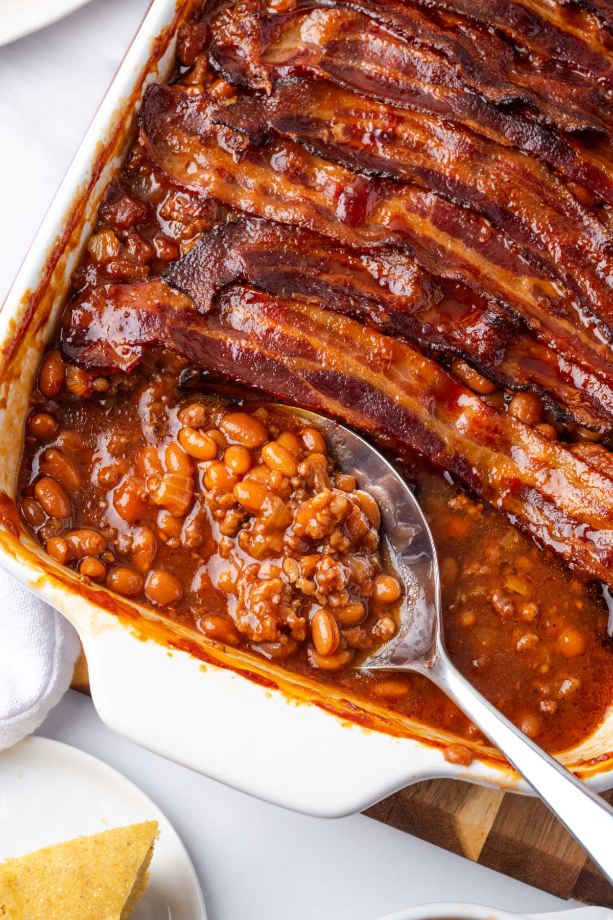Spoon in baked beans with hamburger in white casserole dish, topped with bacon.