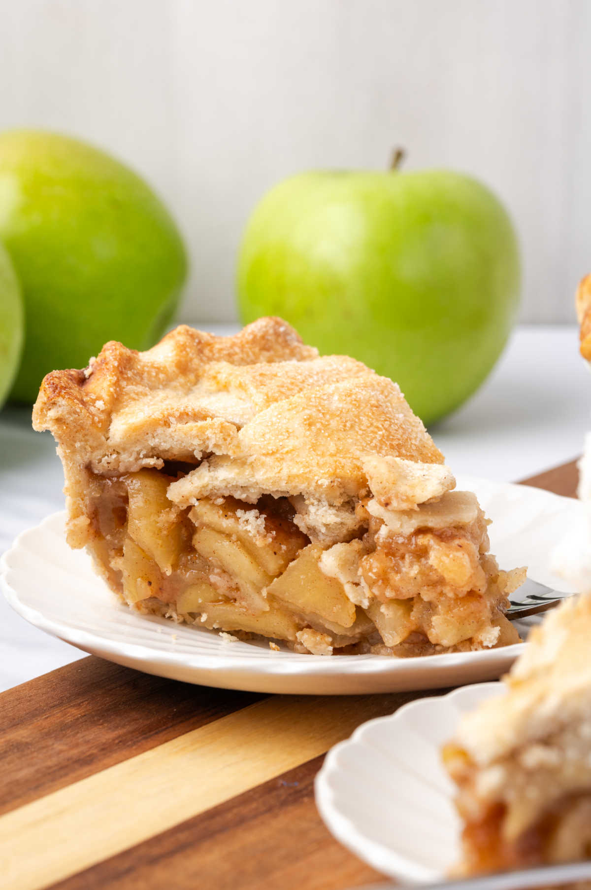 Slice of apple pie on small white plate, green apples in background.