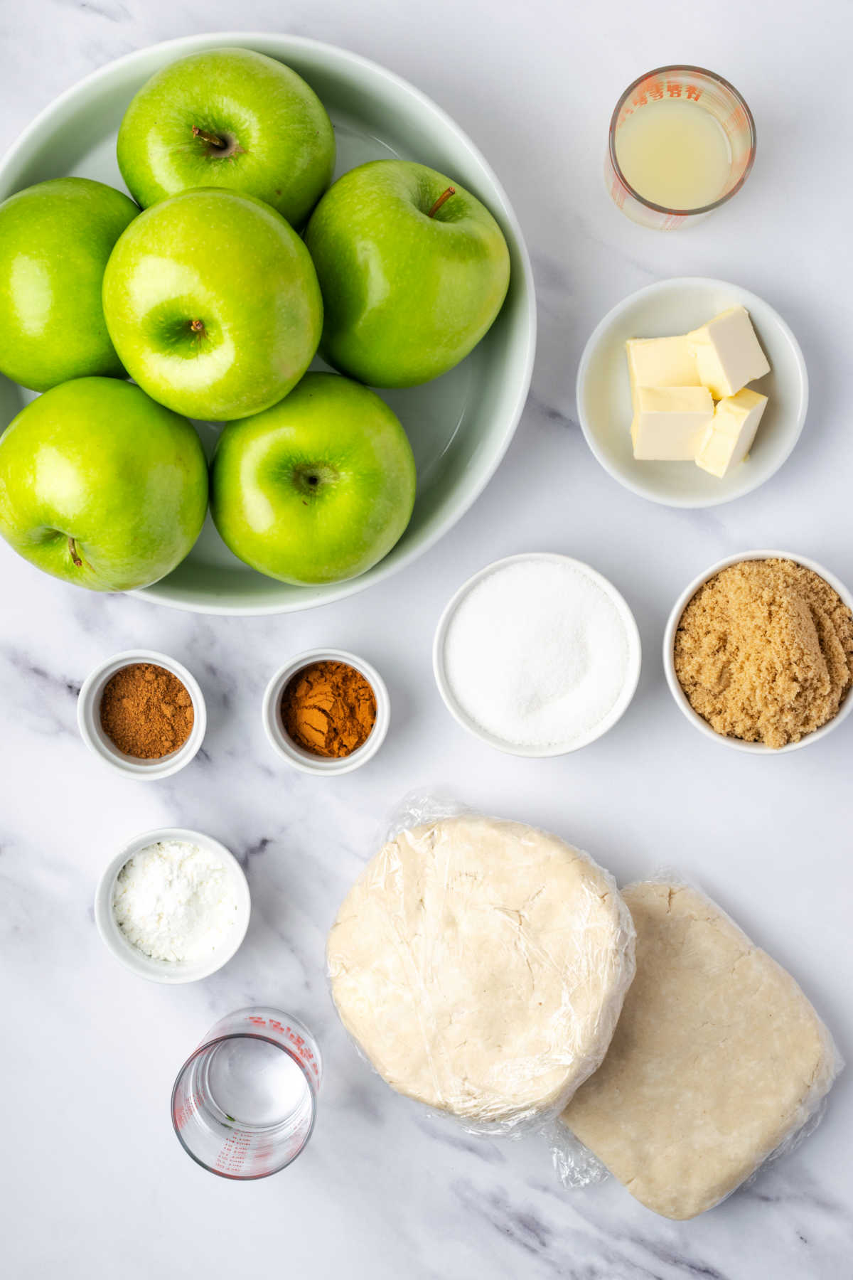Ingredients for apple pie with Granny Smith apples, ingredients in small white bowls on white marble.