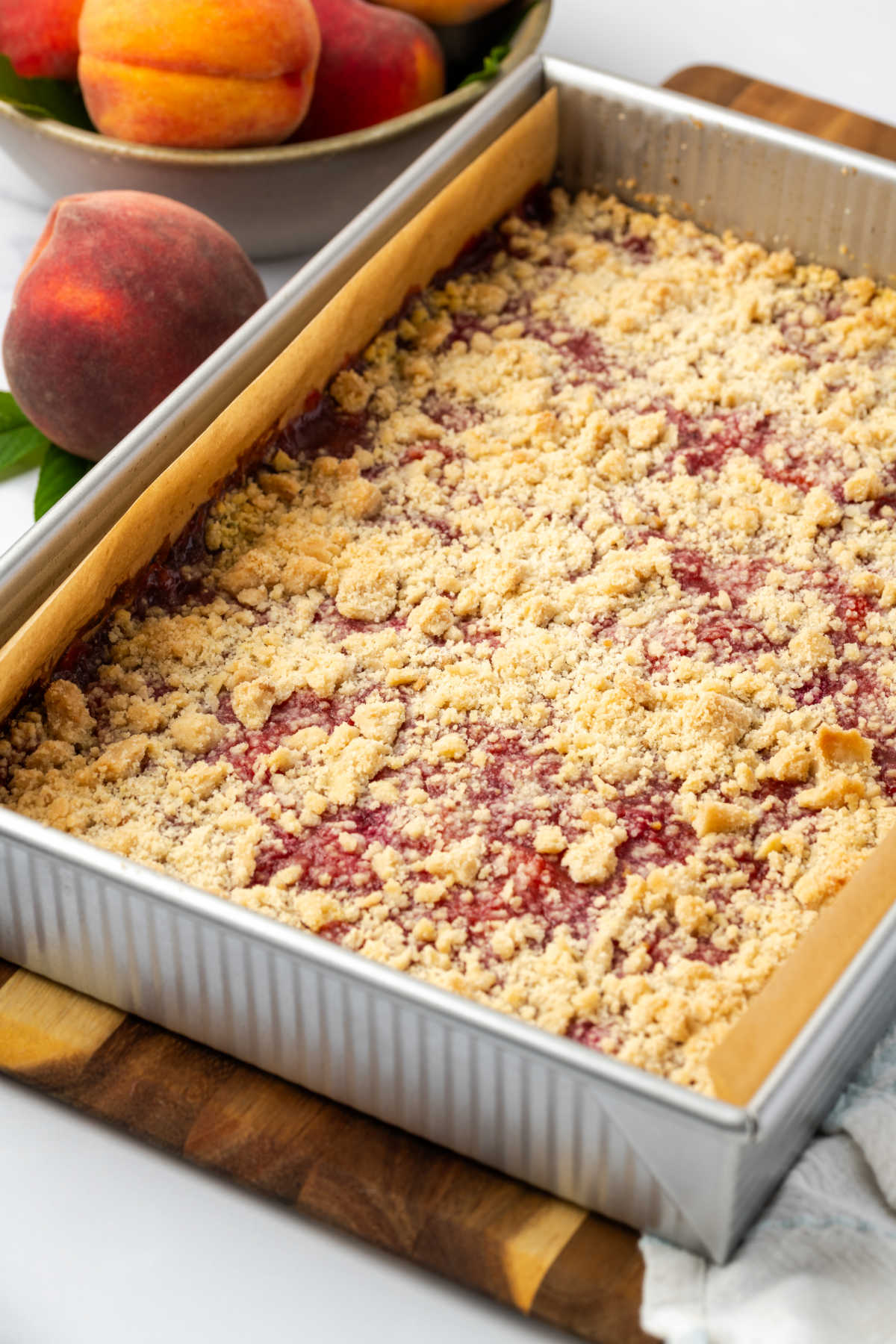 Peach crumb bars in baking pan, fresh peaches in background.