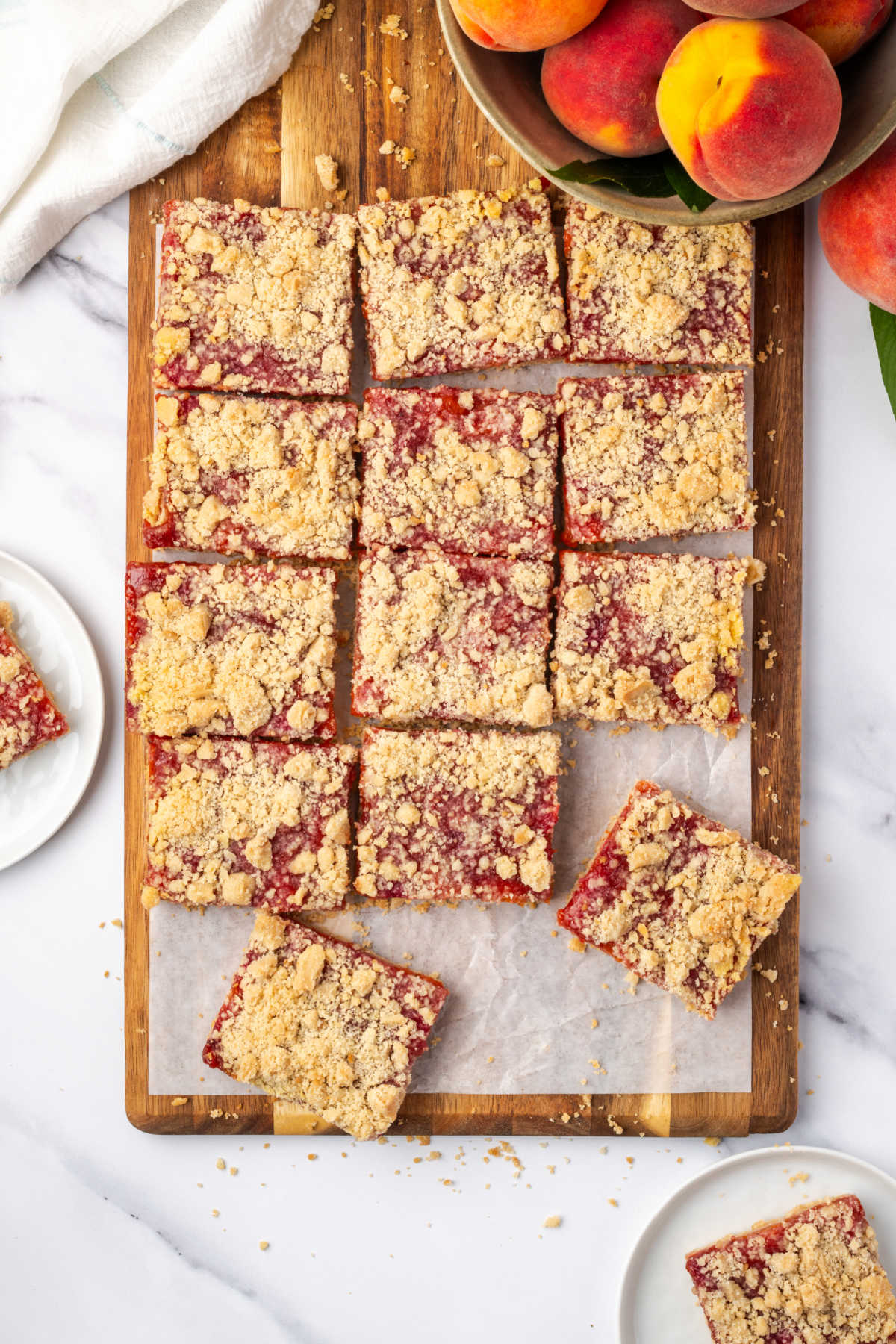 Crumb bars cut into squares on wooden cutting board, fresh peaches in bowl.