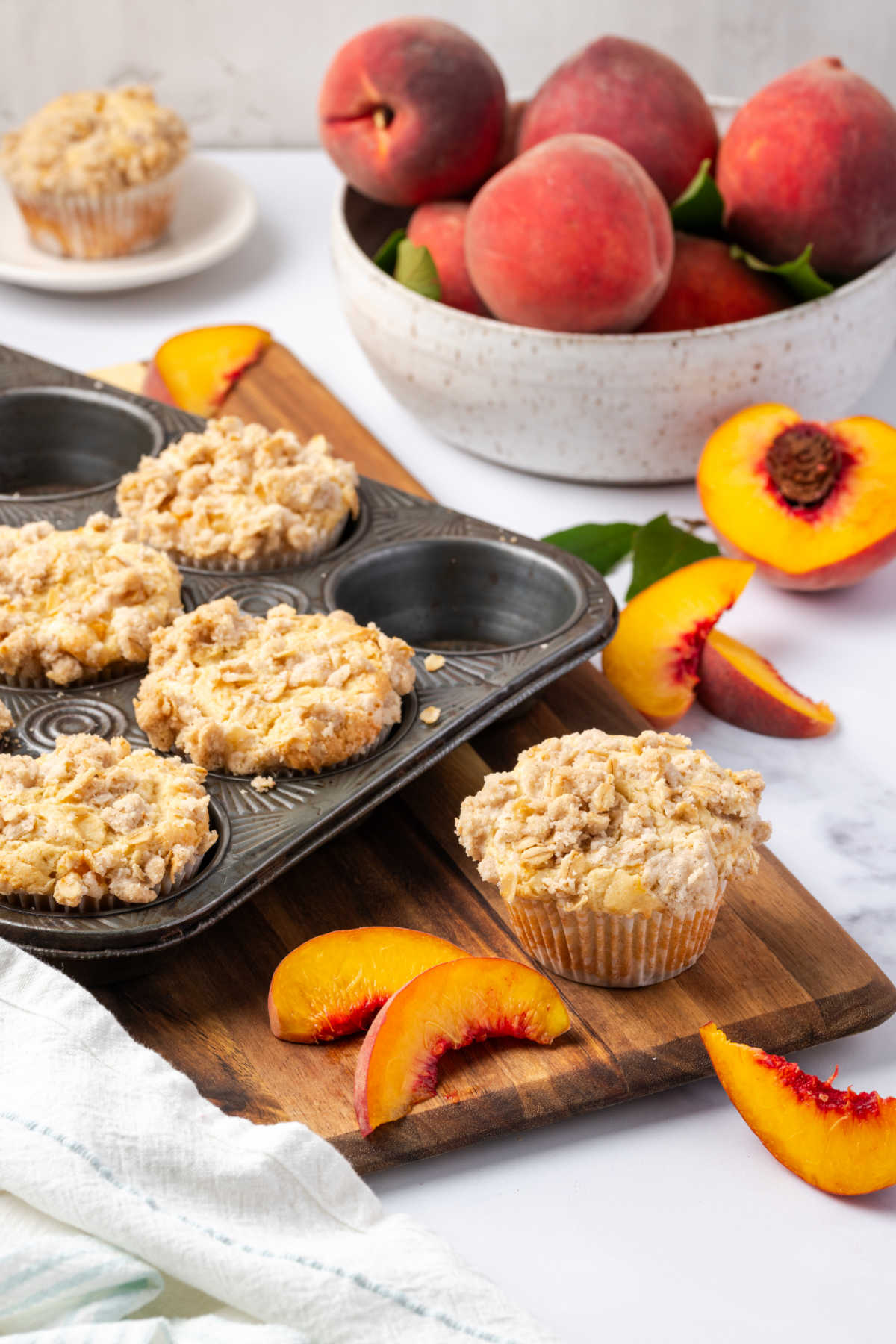 Muffins with crumb topping in metal muffin tin, fresh peaches in background.